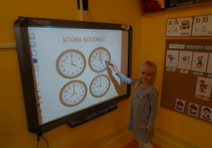 Dziewczynka stoi przy tablicy interaktywnej na której są cztery tarcze zegarów, wskaźnikiem wskazuje na jeden z nich i odczytuje godzinę 5.